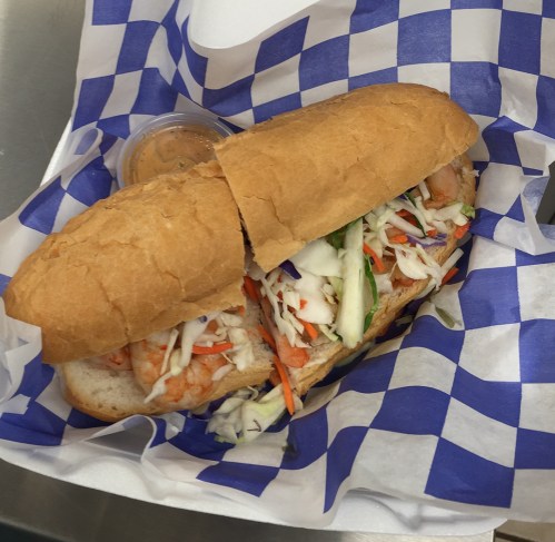 A sandwich filled with shrimp and coleslaw, served in a blue and white checkered paper basket with a dipping sauce.
