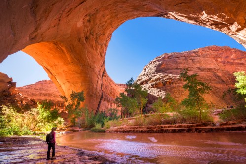 A person stands by a river under a large rock arch, surrounded by vibrant red rock formations and greenery.
