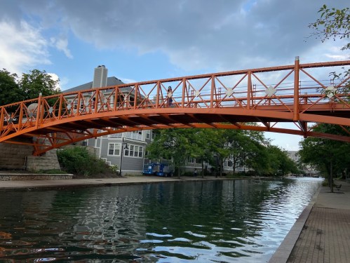 An orange pedestrian bridge spans a calm canal, surrounded by trees and buildings under a cloudy sky.