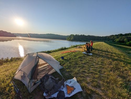 A serene lakeside scene at sunrise with tents set up and two people preparing for the day.