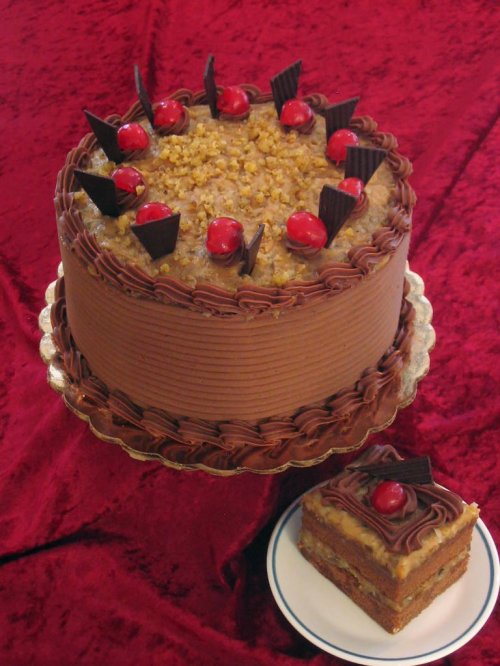 A round chocolate cake topped with cherries, nuts, and chocolate decorations, alongside a slice on a plate.
