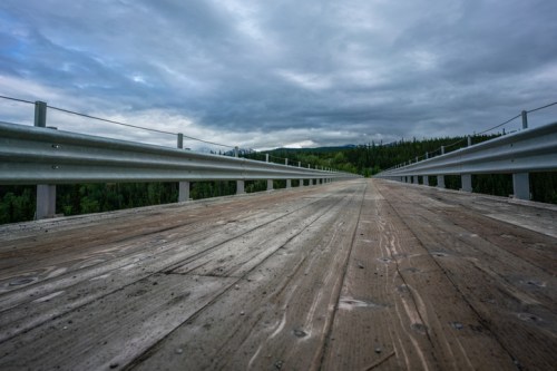 A wooden bridge stretches into the distance under a cloudy sky, flanked by green trees on either side.