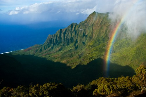 A vibrant rainbow arcs over lush green mountains, with a view of the ocean in the background under a partly cloudy sky.