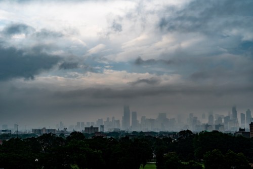 A city skyline shrouded in mist and clouds, with tall buildings partially obscured against a gray sky.