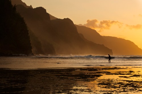 A silhouette of a person walking along a beach at sunset, with mountains and mist in the background.