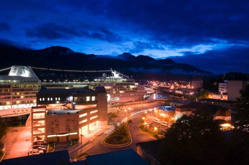 A scenic view of a city at dusk, with buildings, a cruise ship, and mountains under a blue sky.