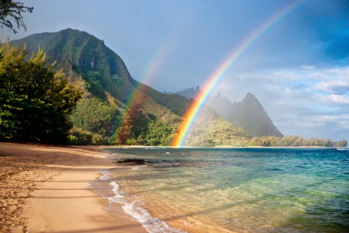 A serene beach scene with gentle waves, lush green mountains, and vibrant double rainbows in a clear sky.
