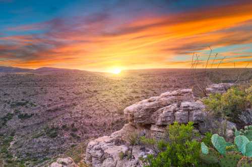 A vibrant sunset over a rugged landscape, featuring rocky terrain and desert vegetation under a colorful sky.