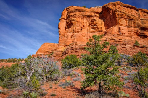 A vibrant red rock formation rises against a blue sky, surrounded by sparse vegetation and desert landscape.