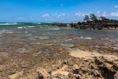 A serene coastal scene with rocky shorelines, clear water, and gentle waves under a blue sky with scattered clouds.