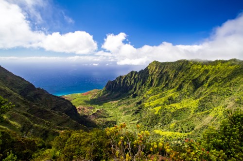 A panoramic view of lush green mountains leading to a blue ocean under a partly cloudy sky.
