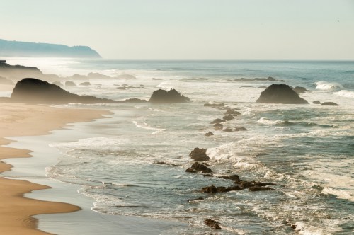 A serene beach scene with gentle waves, rocky formations, and a misty coastline under a clear sky.
