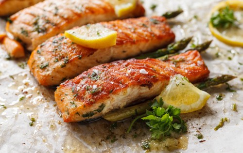 Grilled salmon fillets garnished with lemon slices and fresh herbs, served with asparagus on parchment paper.