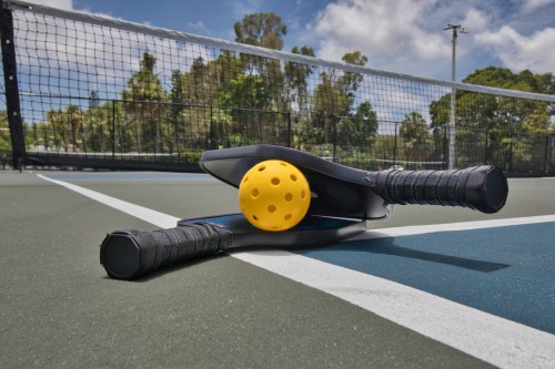A pickleball paddle and a yellow ball resting on a tennis court with a net in the background.