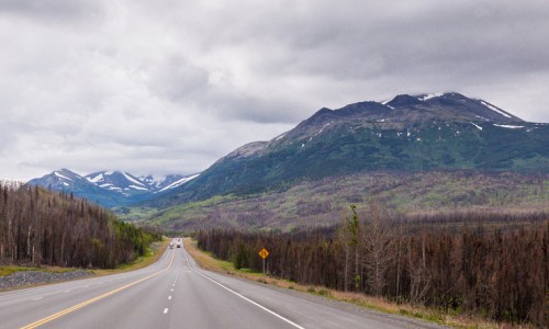 A scenic highway stretches through a mountainous landscape with green hills and patches of snow under a cloudy sky.