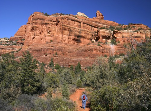 A hiker walks along a trail towards a towering red rock formation under a clear blue sky.