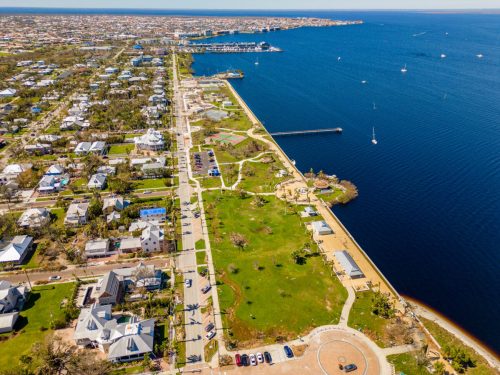 Aerial view of a coastal town with waterfront, parks, and residential areas along the shore. Clear blue water in the background.