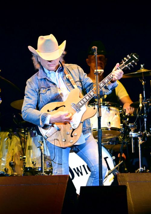 A musician in a cowboy hat plays guitar on stage, with a drummer visible in the background.