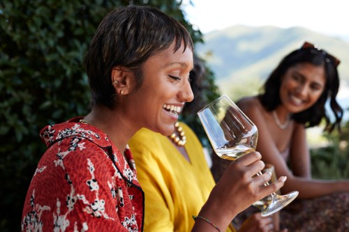 A woman smiles while holding a glass of wine, with two friends enjoying the moment in a scenic outdoor setting.