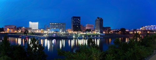 Panoramic view of a city skyline at dusk, reflecting on a river with illuminated buildings and a bridge in the background.