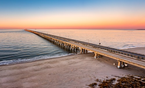 A long pier extends into calm waters at sunset, with soft colors in the sky and sandy beach in the foreground.