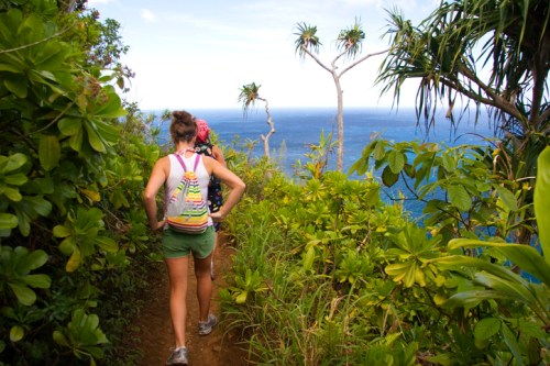 Two hikers walking along a lush, green trail with a view of the ocean in the background.