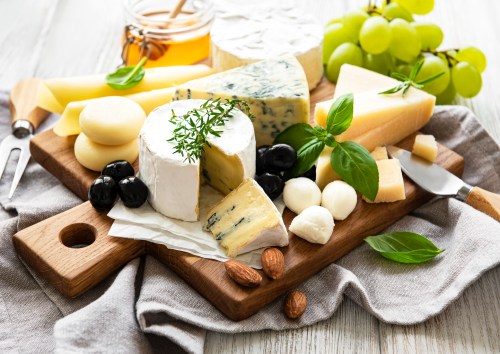 A wooden cheese board featuring various cheeses, olives, grapes, nuts, and herbs, with honey in a jar nearby.