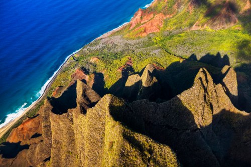 Aerial view of rugged cliffs meeting a vibrant coastline and deep blue ocean, surrounded by lush greenery.