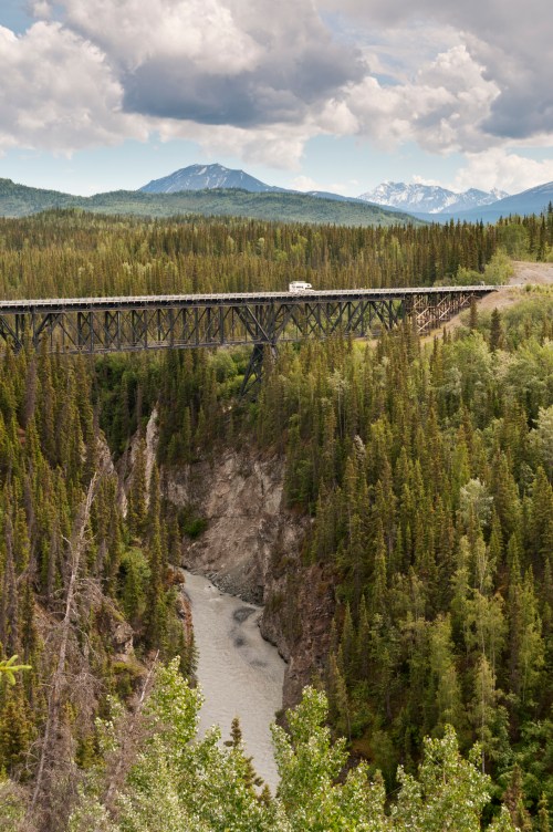 A bridge spans a deep canyon surrounded by lush forests and mountains under a cloudy sky.