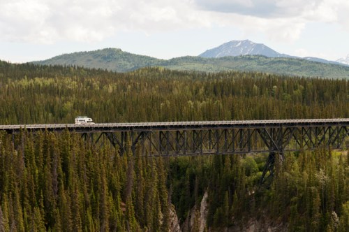 A vehicle crosses a tall wooden bridge surrounded by lush green forests and distant mountains under a cloudy sky.