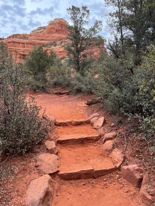 A winding dirt path with stone steps leads through lush greenery towards a red rock formation under a cloudy sky.