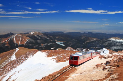 A red train travels along snowy tracks on a mountain, with a panoramic view of valleys and lakes in the background.