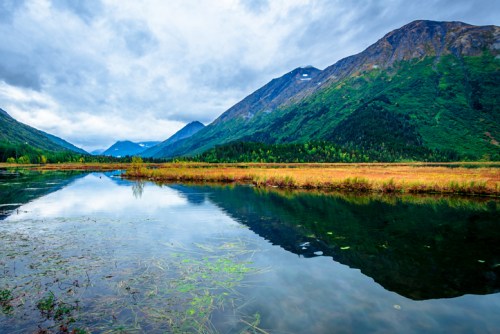 A serene landscape featuring a calm lake reflecting mountains and lush greenery under a cloudy sky.