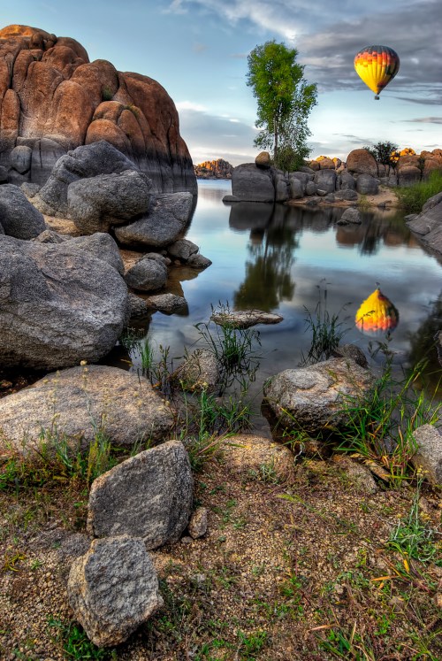A serene landscape featuring rocky shores, a calm lake, and a colorful hot air balloon reflecting in the water.