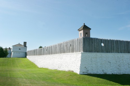 A historic fort with a wooden palisade and lookout tower, set against a clear blue sky and green grass.