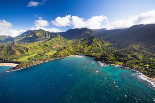 Aerial view of lush green mountains meeting a turquoise ocean under a bright blue sky with scattered clouds.