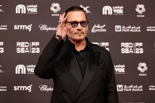 A man in a black suit and sunglasses waves at the camera on a red carpet with event branding in the background.