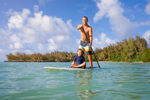 A man and a boy paddleboarding together in clear water, surrounded by lush greenery and a blue sky.