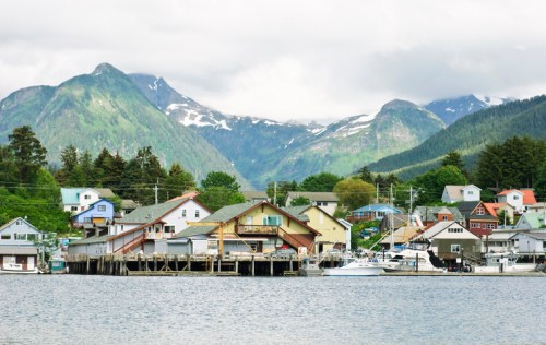 A scenic view of a waterfront town with colorful buildings, boats, and mountains in the background under a cloudy sky.