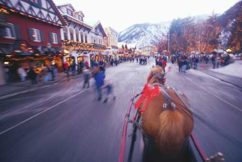 A horse-drawn carriage rides through a festive, snow-covered town with holiday lights and bustling crowds.