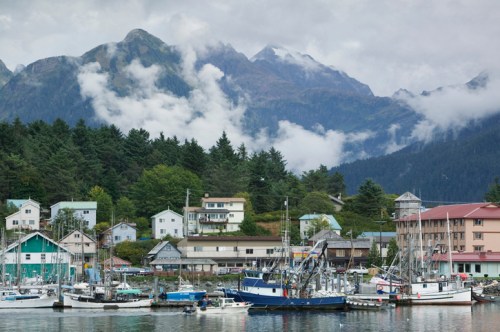 A scenic harbor with fishing boats, surrounded by lush trees and mountains under a cloudy sky.