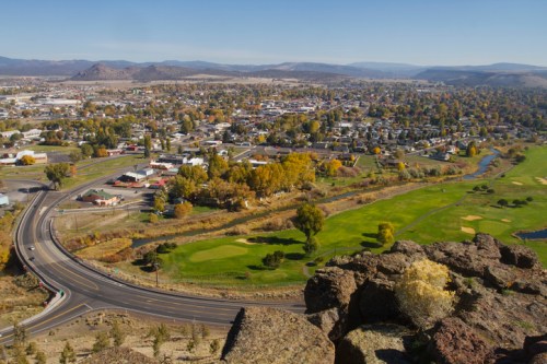 Aerial view of a small town with green fields, winding roads, and mountains in the background under a clear blue sky.