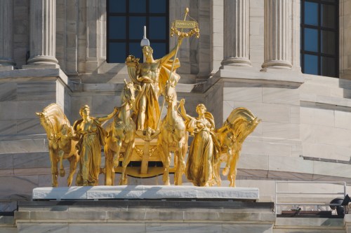 Golden statue of a chariot with horses and figures, set against a marble building backdrop.