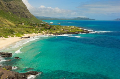 A scenic view of a turquoise ocean meeting a sandy beach, framed by lush green hills under a partly cloudy sky.