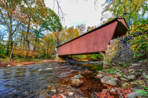 A red covered bridge spans a stream, surrounded by autumn foliage and rocky banks.