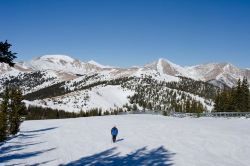 A skier glides across a snowy landscape with mountains and clear blue skies in the background.