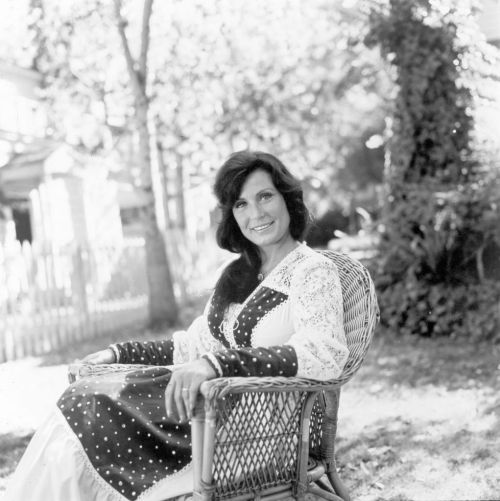 A woman in a polka dot dress sits in a wicker chair outdoors, smiling in a sunlit garden.