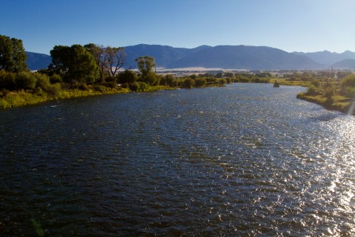 A serene river flows through a landscape of trees and mountains under a clear blue sky.