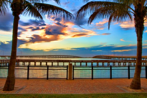 Sunset over a calm waterfront with a wooden pier, framed by palm trees and colorful clouds in the sky.