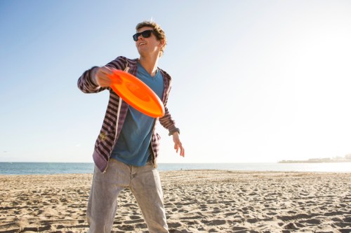 A young man in sunglasses throws an orange frisbee on a sunny beach. The ocean is visible in the background.